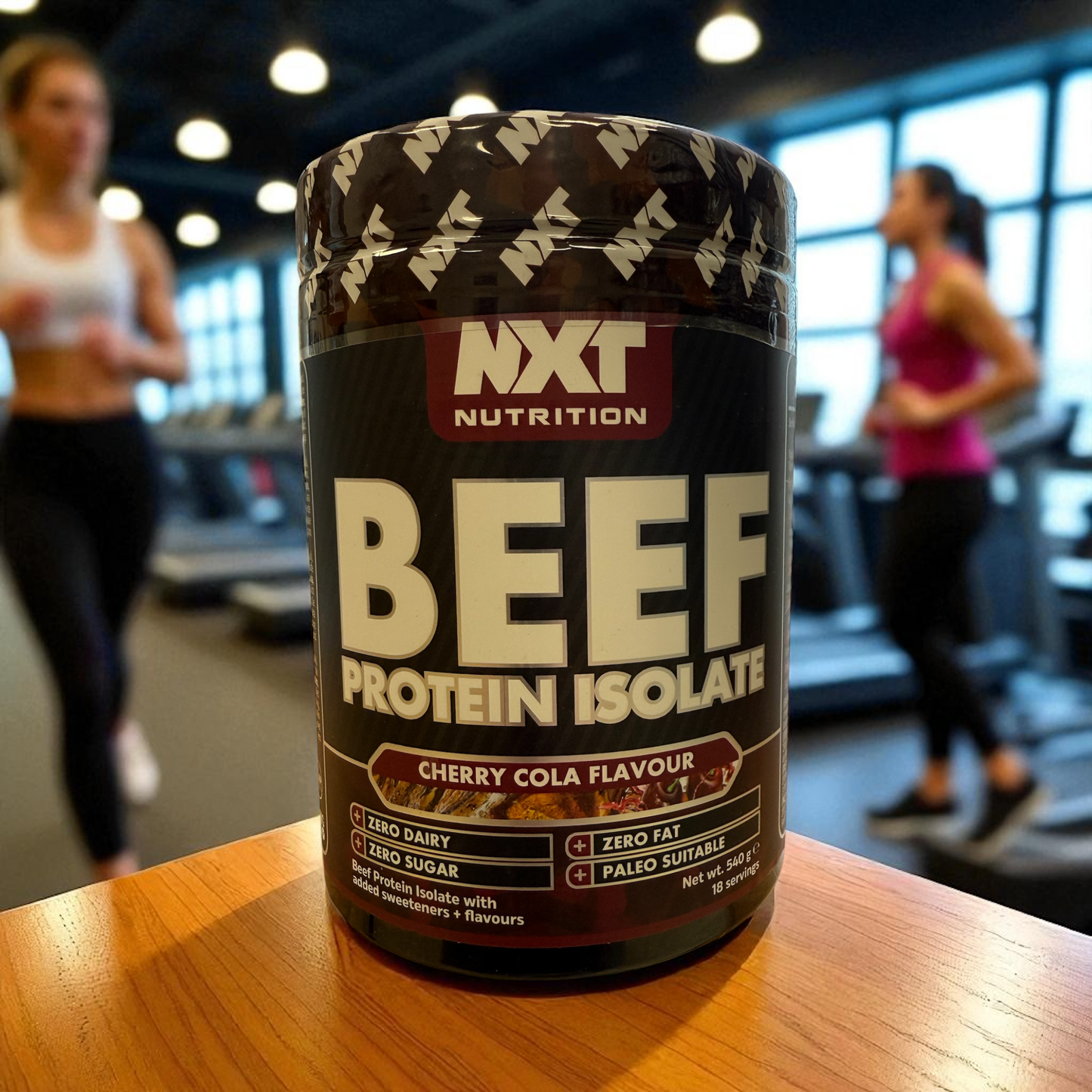 NXT Nutrition Beef Protein Isolate container on a table with people exercising in the background, in Cherry cola flavour
