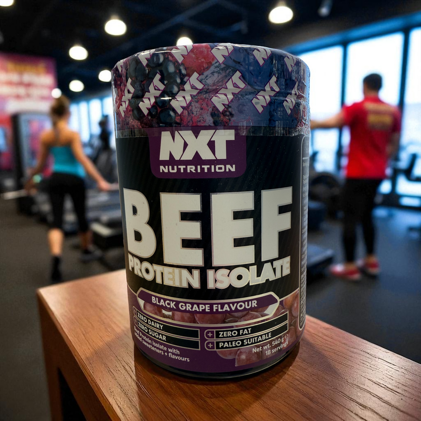 NXT Nutrition Beef Protein Isolate container on a table with people exercising in the background in black grape