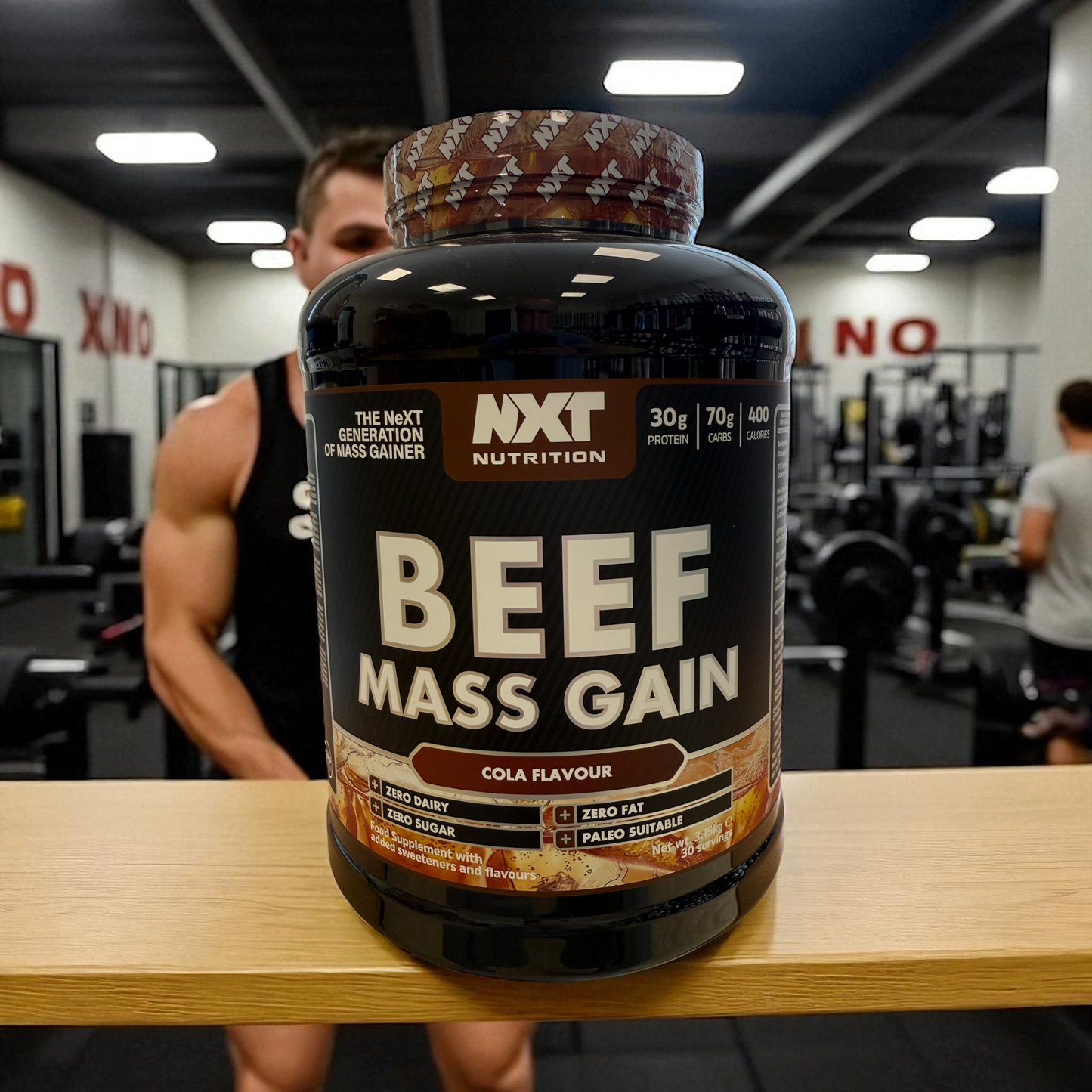 Cola flavour 3kg tub NXT Nutrition Beef Mass Gain supplement bottle on a table with a person in a gym setting.