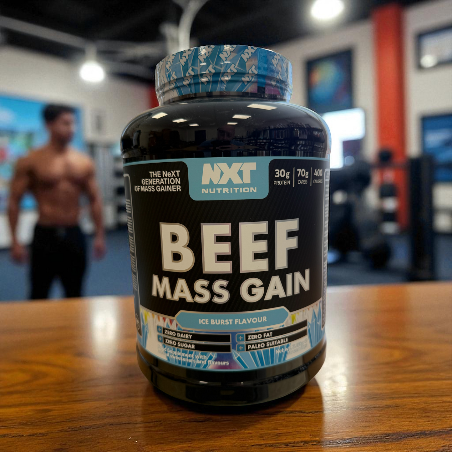 3kg tub of NXT Nutrition Beef Mass Gain supplement in Ice Blast flavour on a wooden table with a blurred gym background.