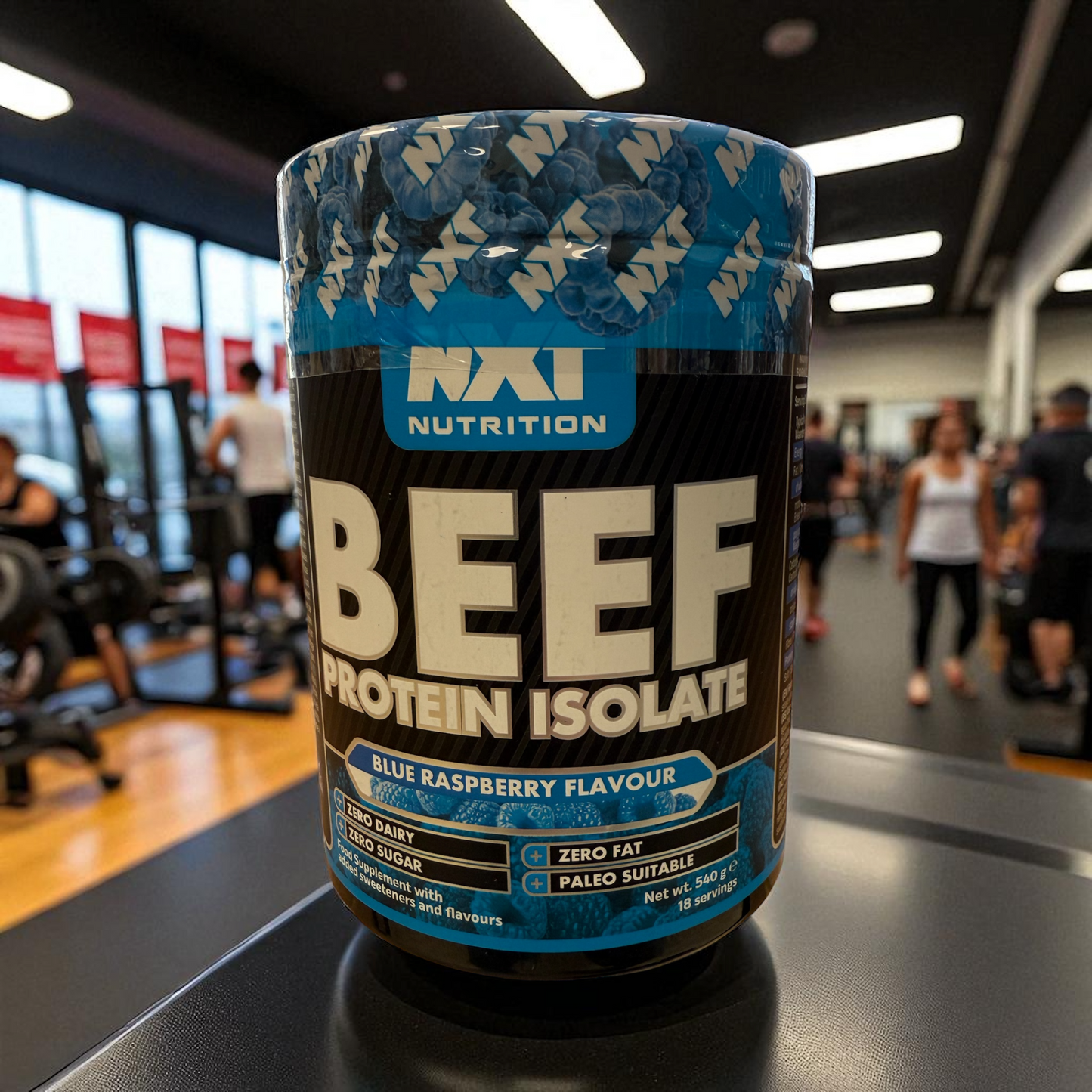 Container of beef protein isolate with Nxt Nutrition branding in a gym setting in blue raspberry flavour