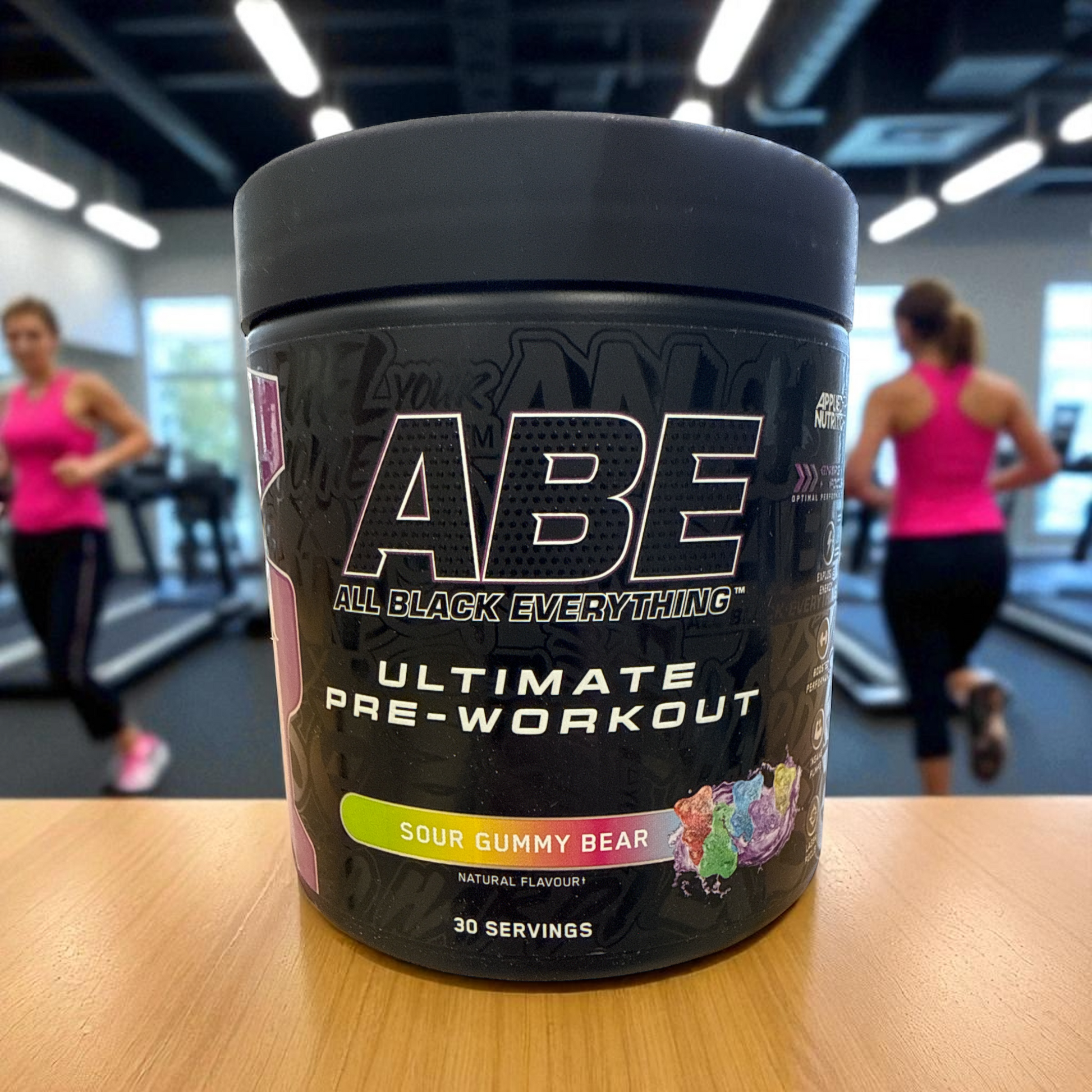 ABE Ultimate Pre-Workout container on a table with people exercising in the background