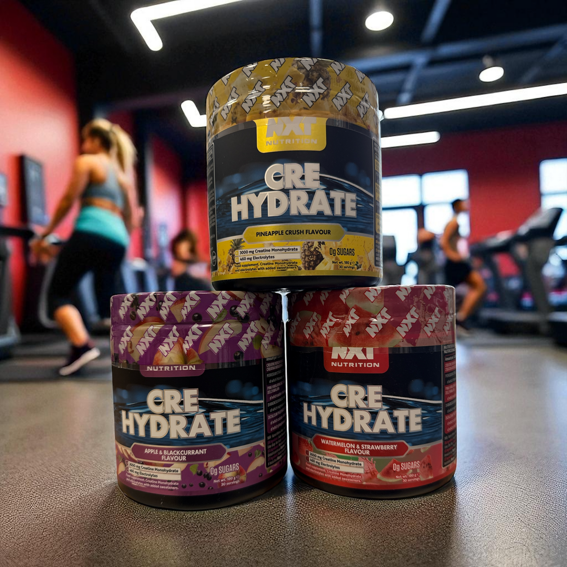 Three containers of Cre Hydrate supplement stacked on a gym floor with people exercising in the background.